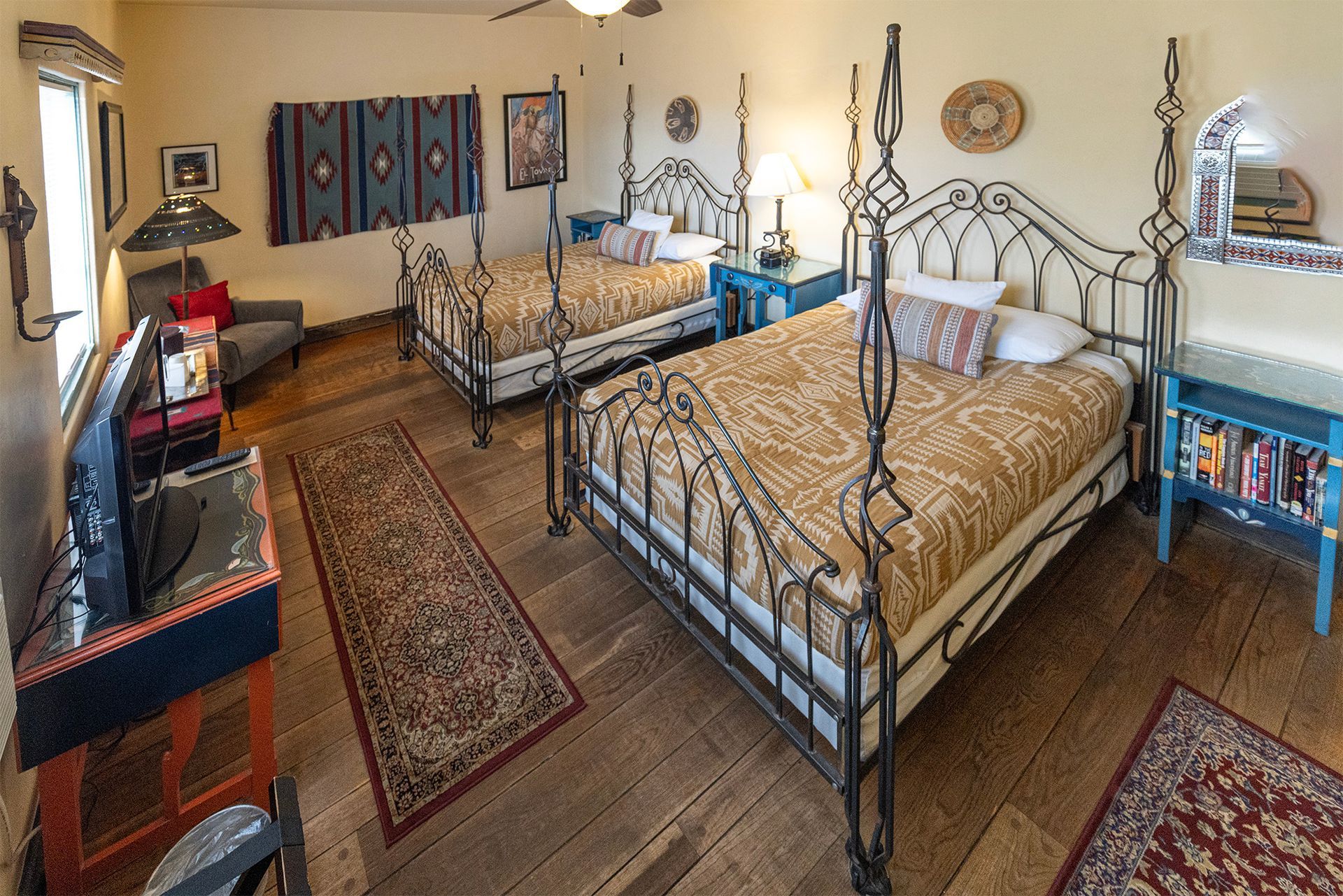 Two beds with decorative metal frames in a beige bedroom, with dark wood floors and colorful decor.