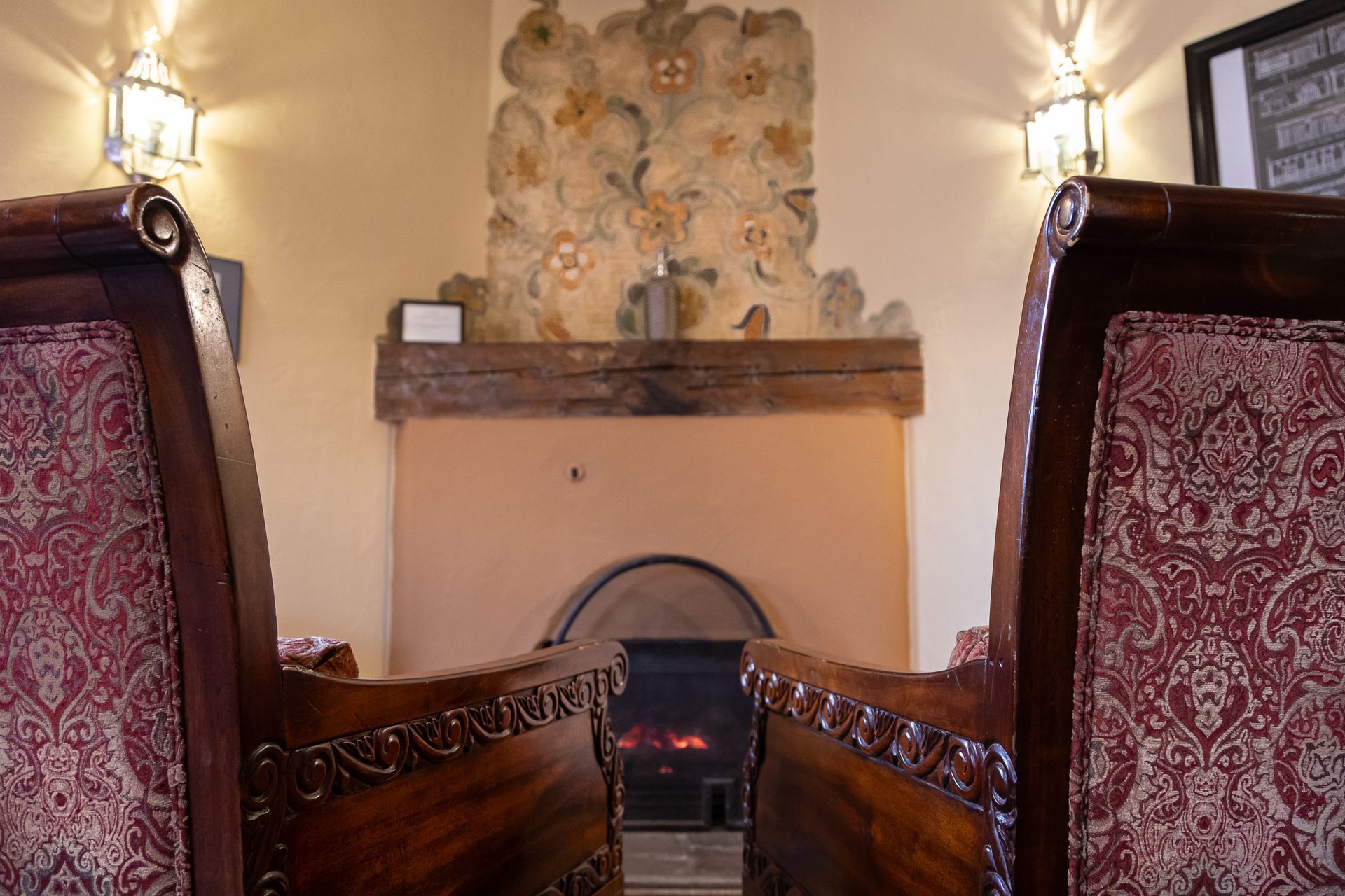 Two ornate chairs frame a fireplace with floral mural, wood mantel, and flickering flames.