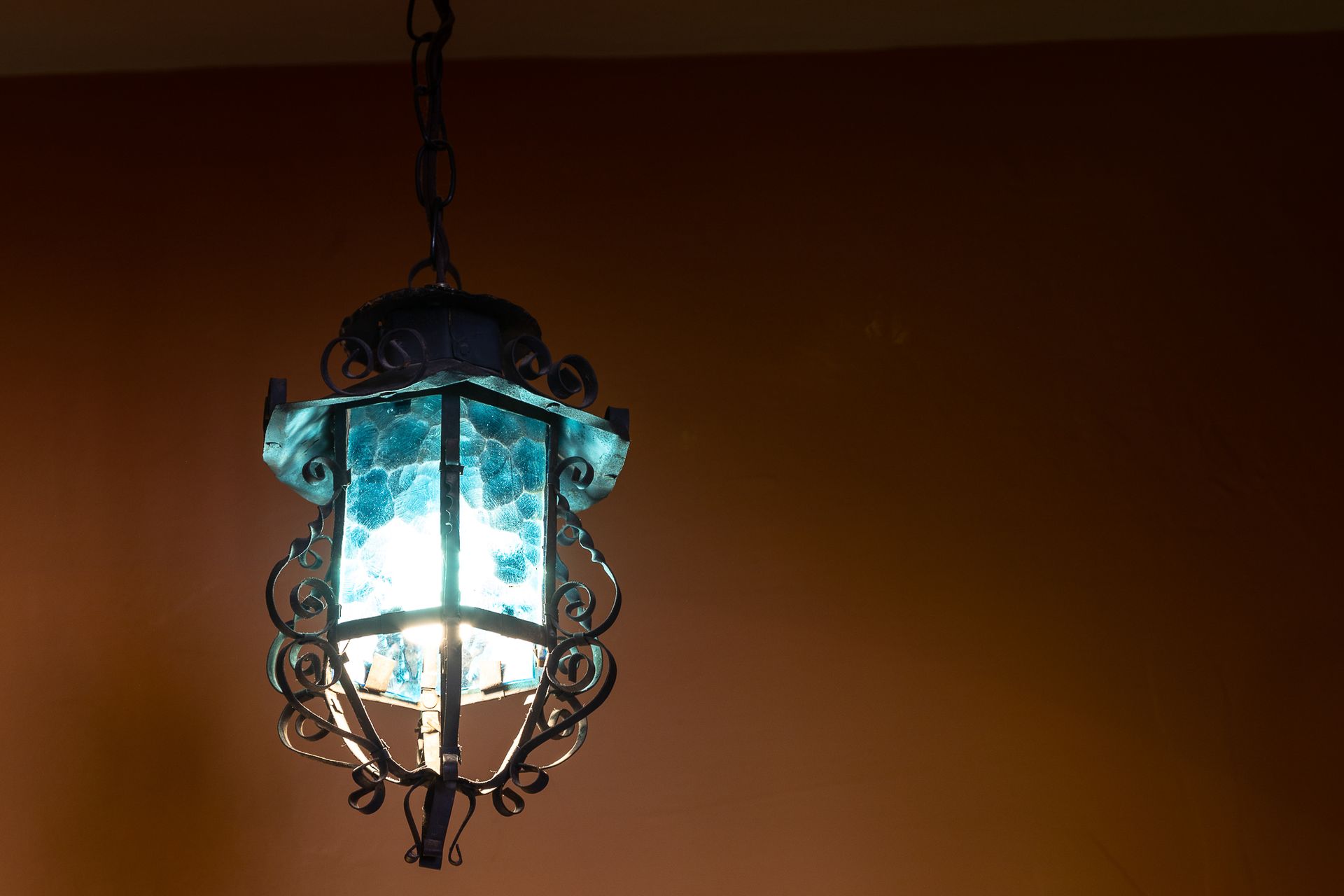 Ornate blue glass and wrought-iron lantern hanging from a chain against a reddish-brown wall.