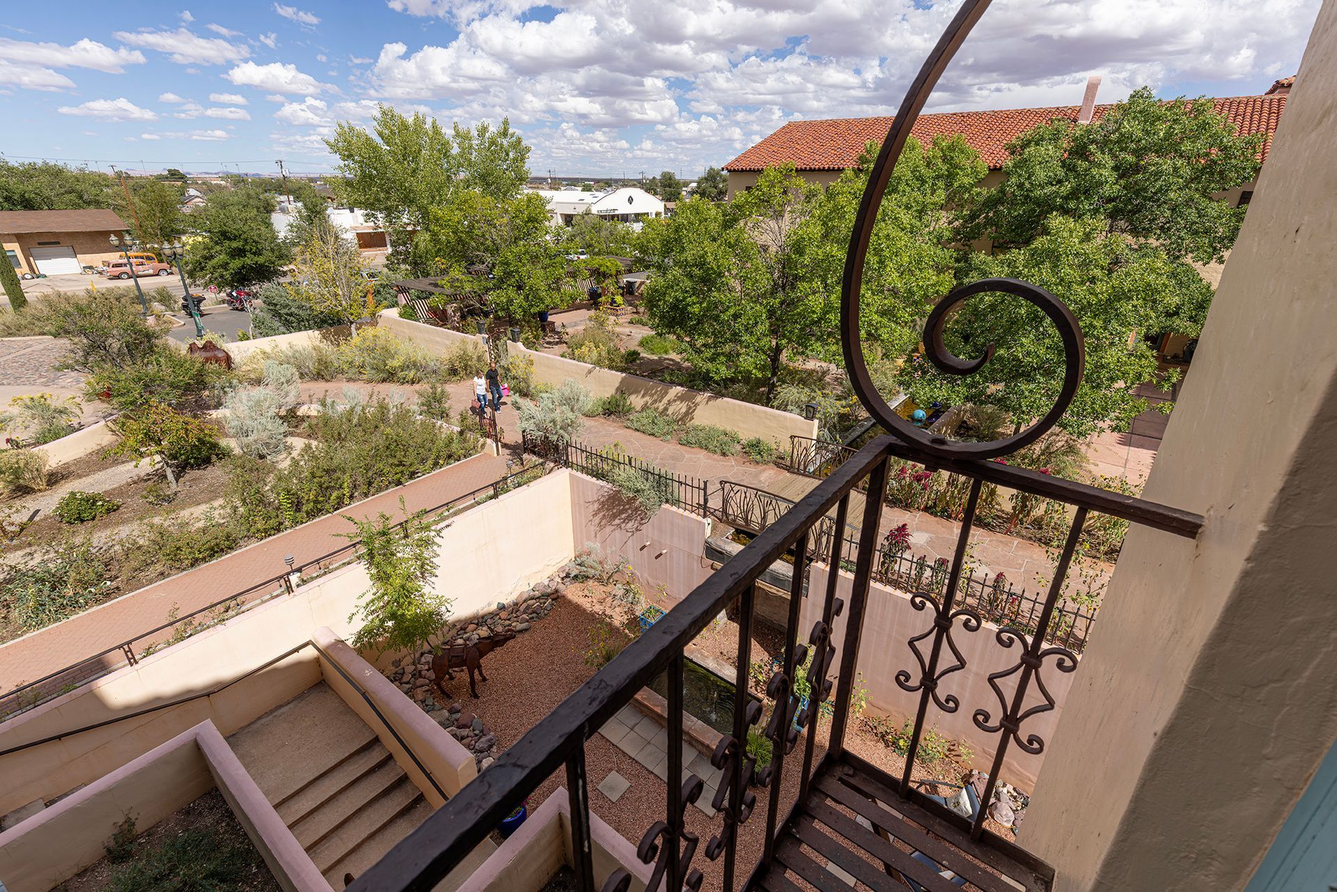 View from a balcony with decorative ironwork, overlooking a garden with stone paths and trees under a cloudy sky.