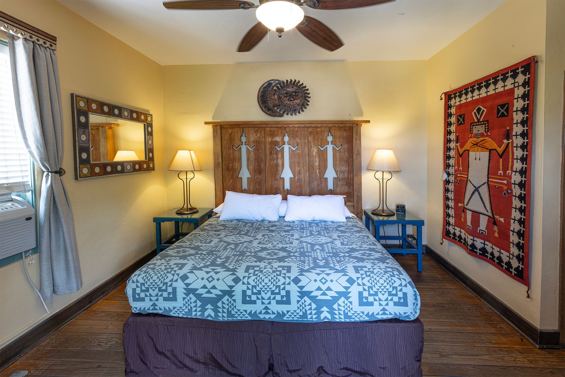 Bedroom with queen bed, wooden headboard, blue side tables, tapestry, mirror, and tan walls.