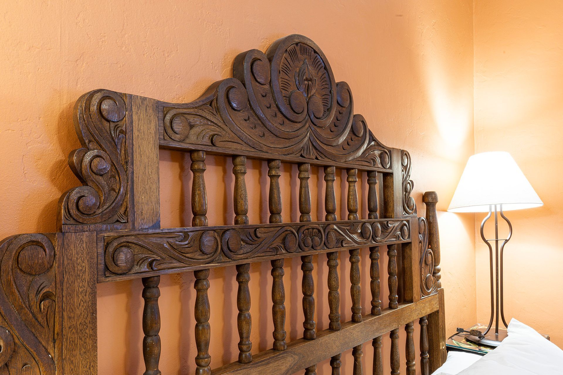 Ornate wooden headboard against an orange wall, beside a lamp on a nightstand.
