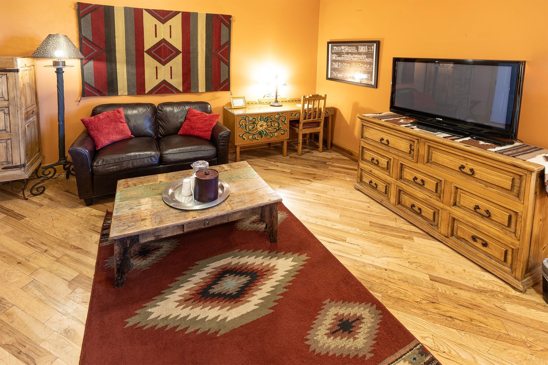 Southwestern-style hotel room: leather couch, red rug, wood furniture, orange walls, TV, decorative rug on wall.