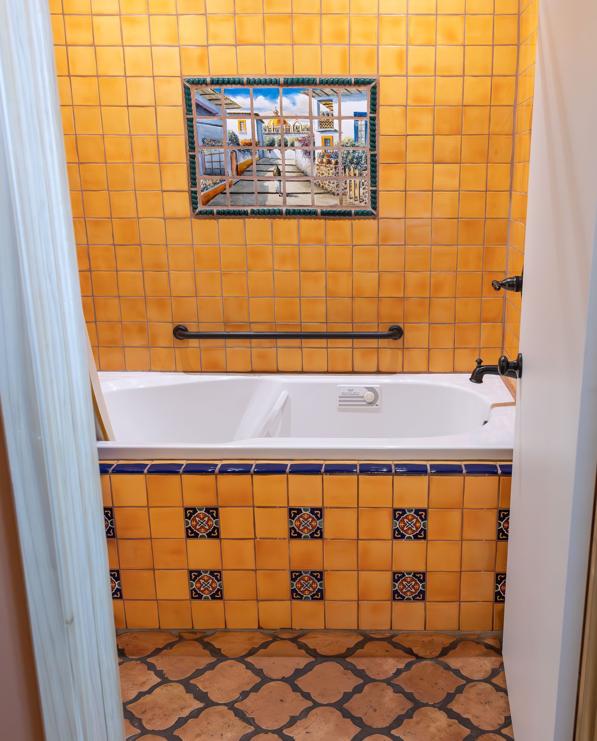 Bathroom with yellow tiled walls, tub, and decorative tile panel. Black handrail.