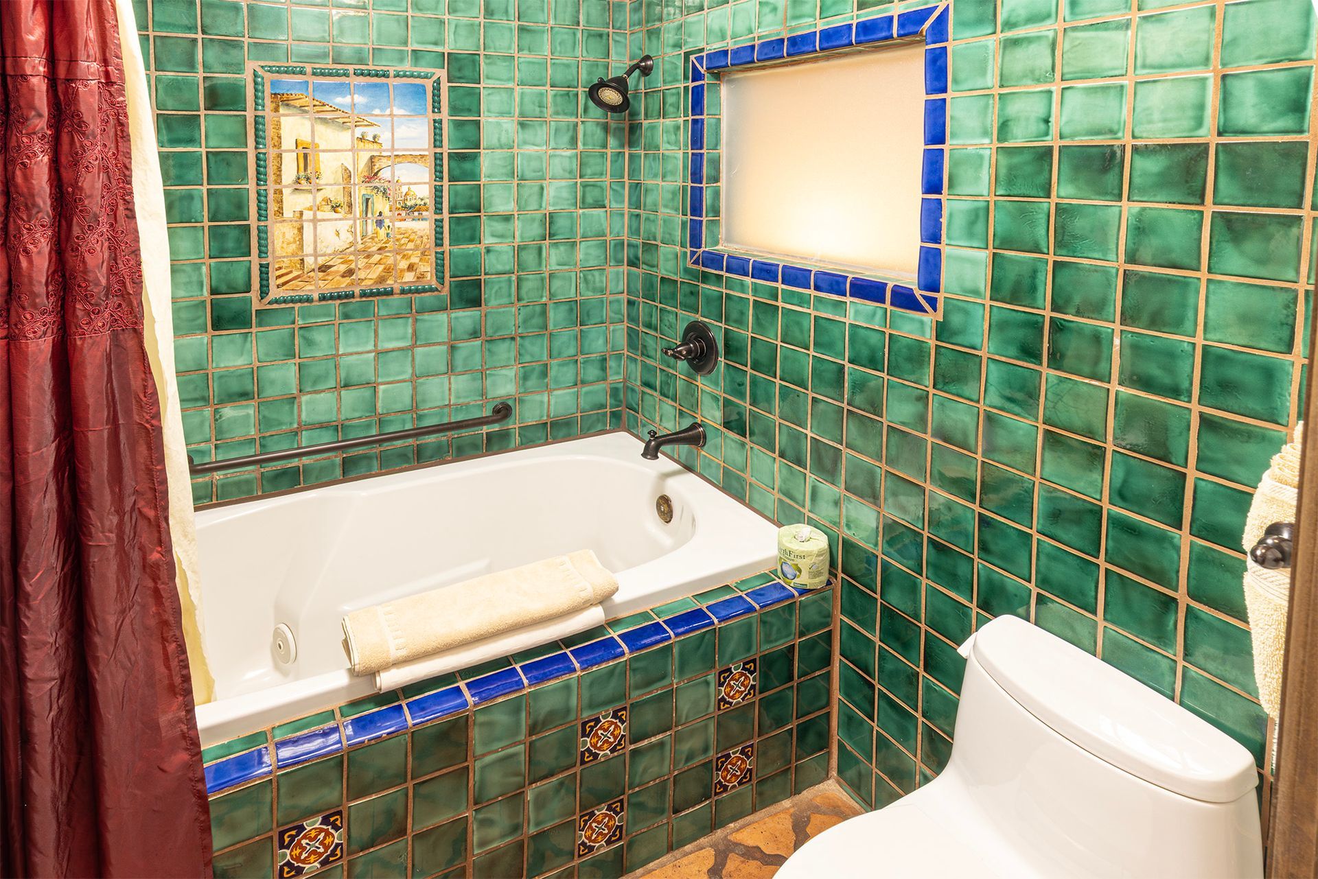 Green tiled bathroom with white tub, toilet, and frosted window, with decorative art and blue accents.