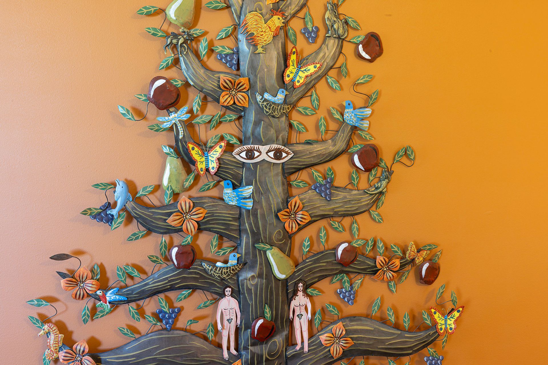 Decorative tree sculpture with fruit, birds, butterflies, and figures of Adam and Eve on an orange wall.