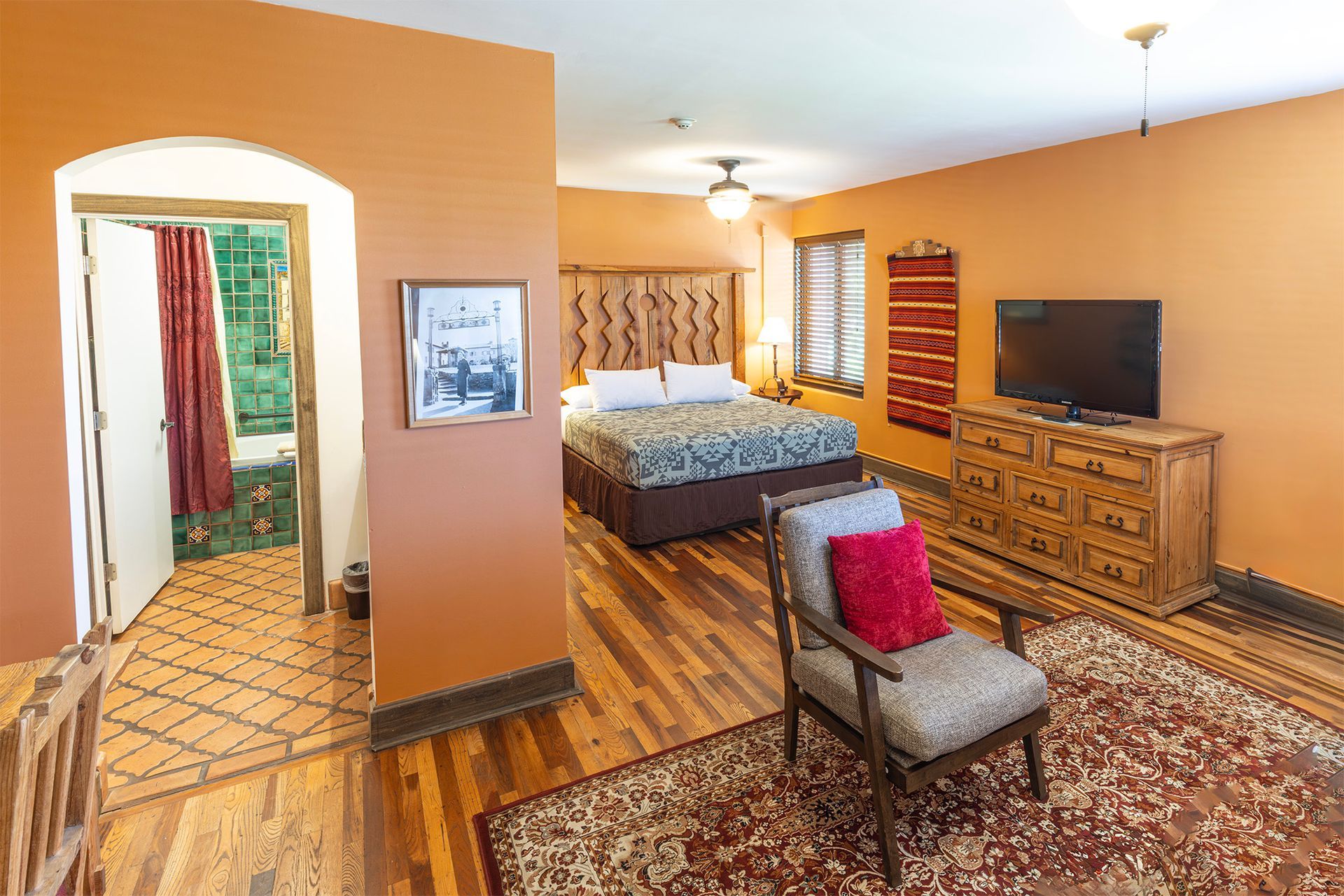 Hotel room with bed, TV, chair, and archway to another room. Orange walls, wooden floor, and rug.