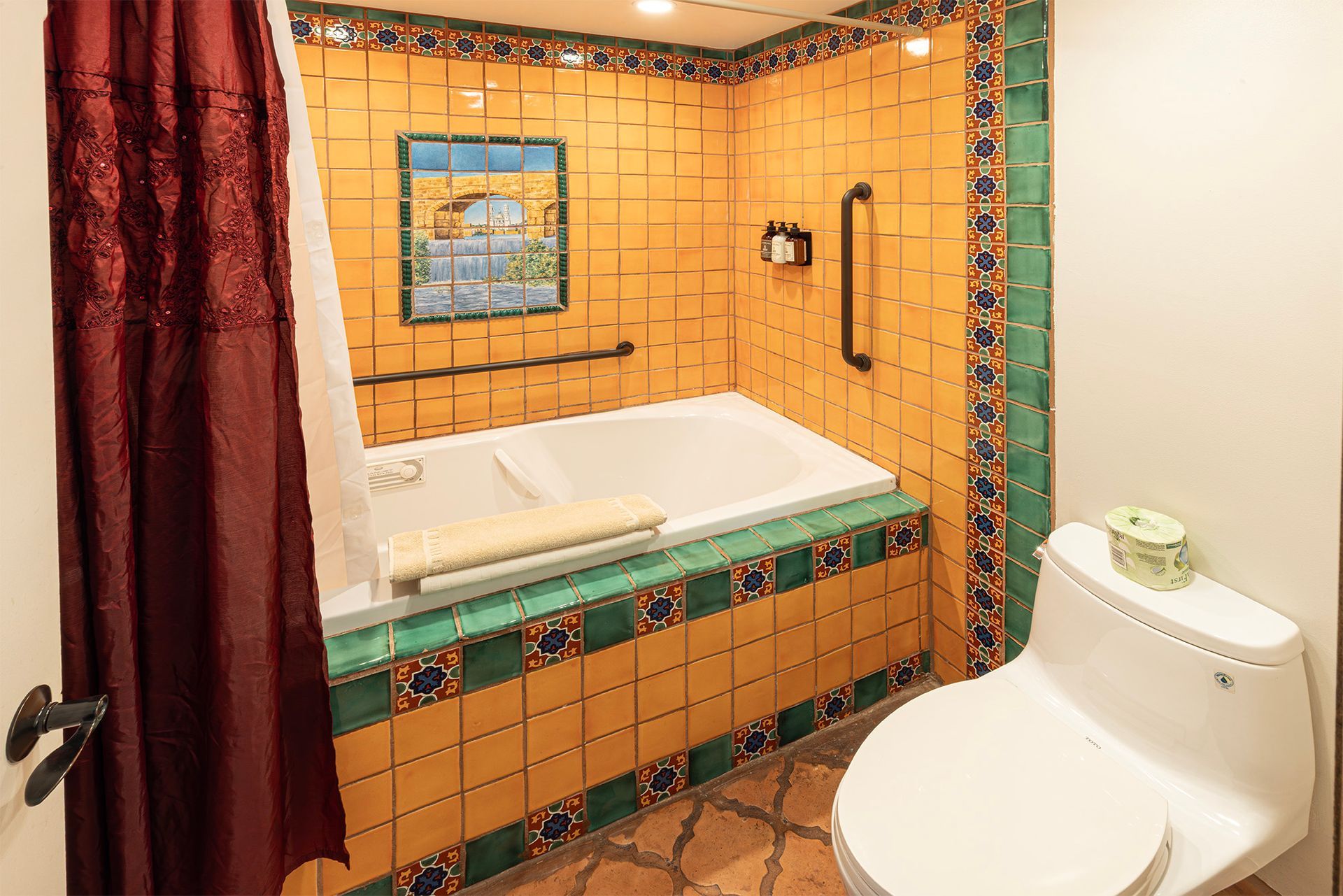Bathroom with yellow tiled walls, tub with handrail, and toilet; southwestern style.