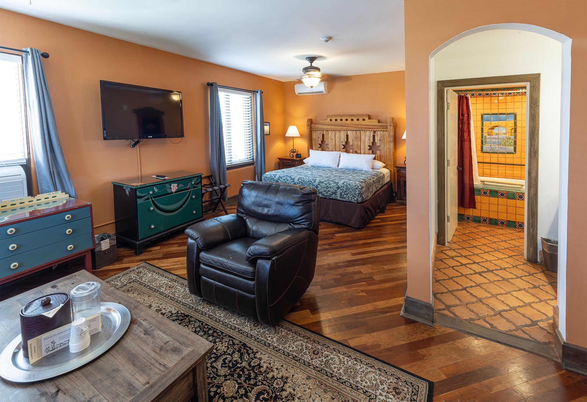 Hotel room with bed, chair, TV, and bathroom entrance. Warm colors and wooden floors.
