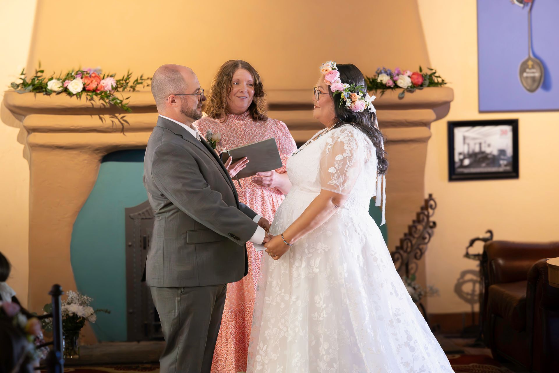Couple holding hands at altar during wedding ceremony. Officiant in pink dress. Floral decorations.