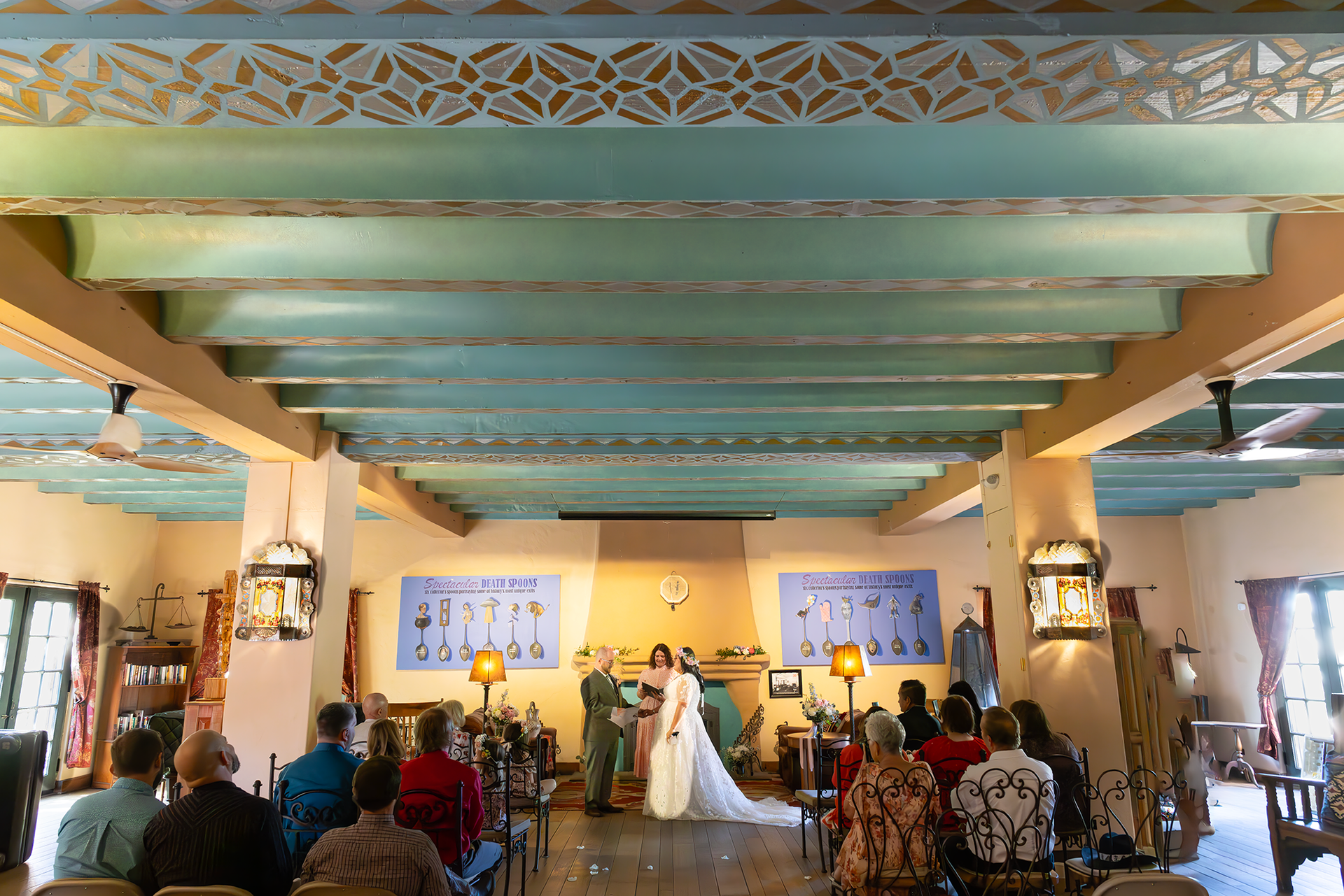 Wedding ceremony inside a building with a light blue ceiling and wooden beams; couple at the altar, guests seated.