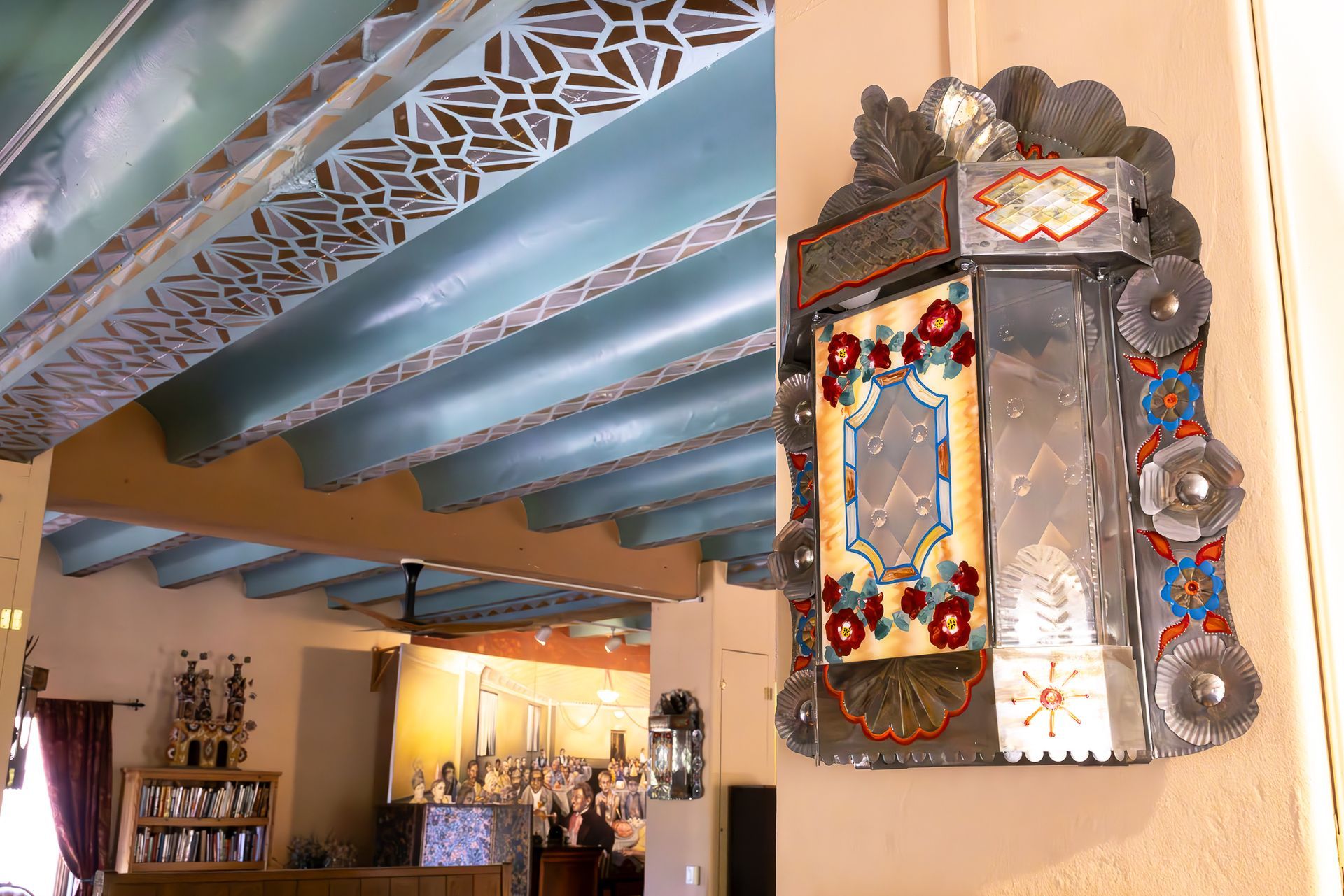 Metalwork plaque on a wall with colorful embellishments, near a room with blue ceiling beams and art.