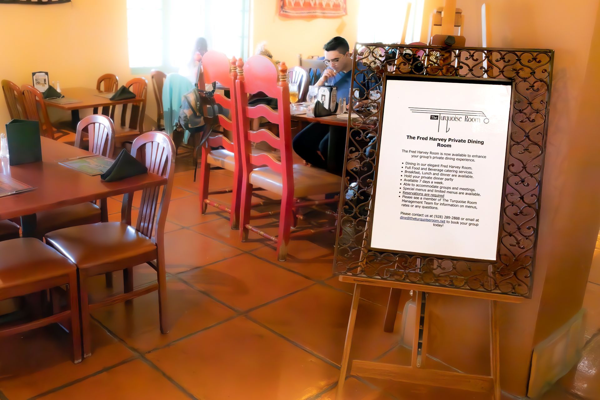 Inside a restaurant, a menu on a decorative easel stands near tables and chairs; a few patrons are seated.