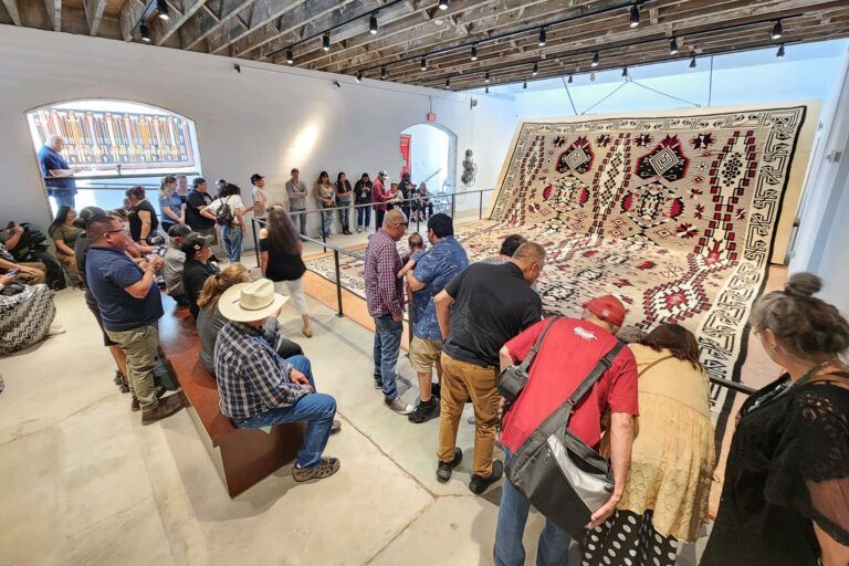 People viewing a large, patterned textile hanging in an art gallery.
