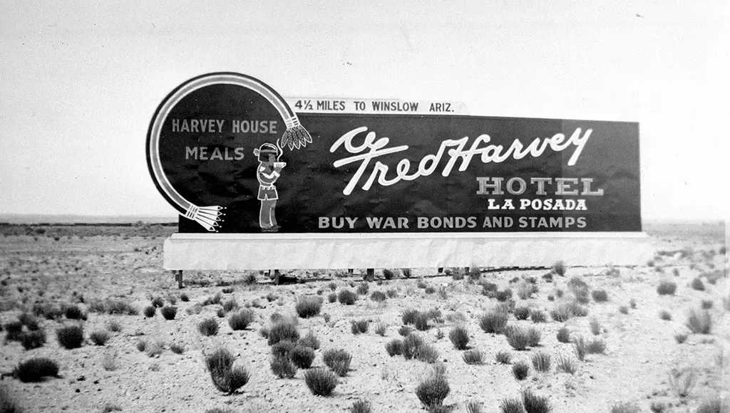 Large sign for Fred Harvey Hotel, La Posada, in desert landscape, promoting meals and war bonds.