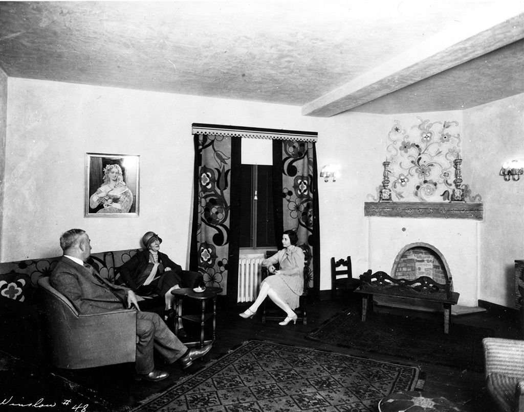 Living room scene: Three people seated on furniture, fireplace, dark curtains, wall art, lamps.