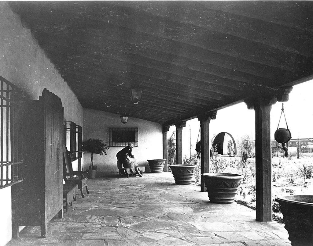 Covered porch with stone floor, wooden posts, potted plants, and two figures.