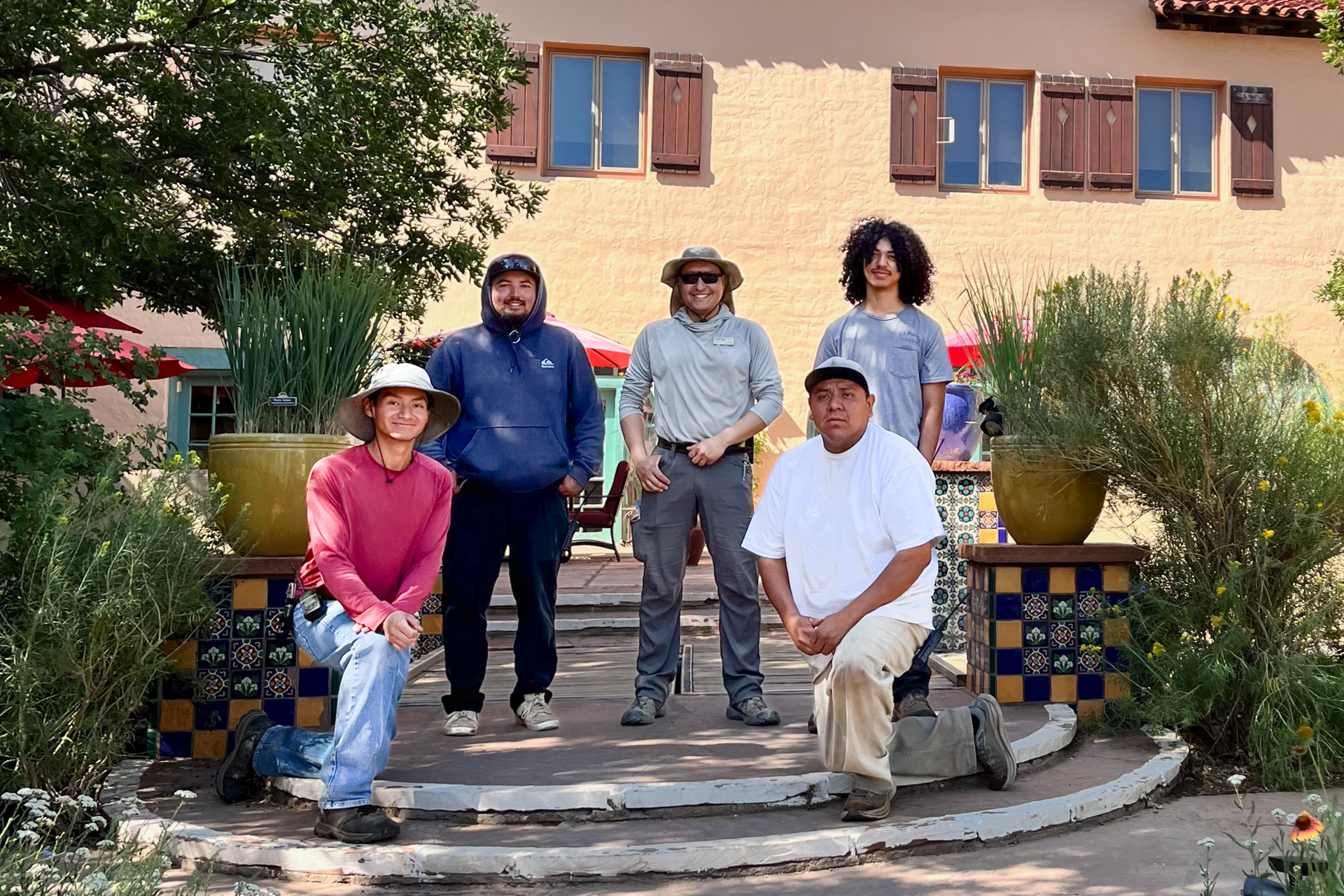 Five people posing on steps outside a building with colorful tile railings and potted plants