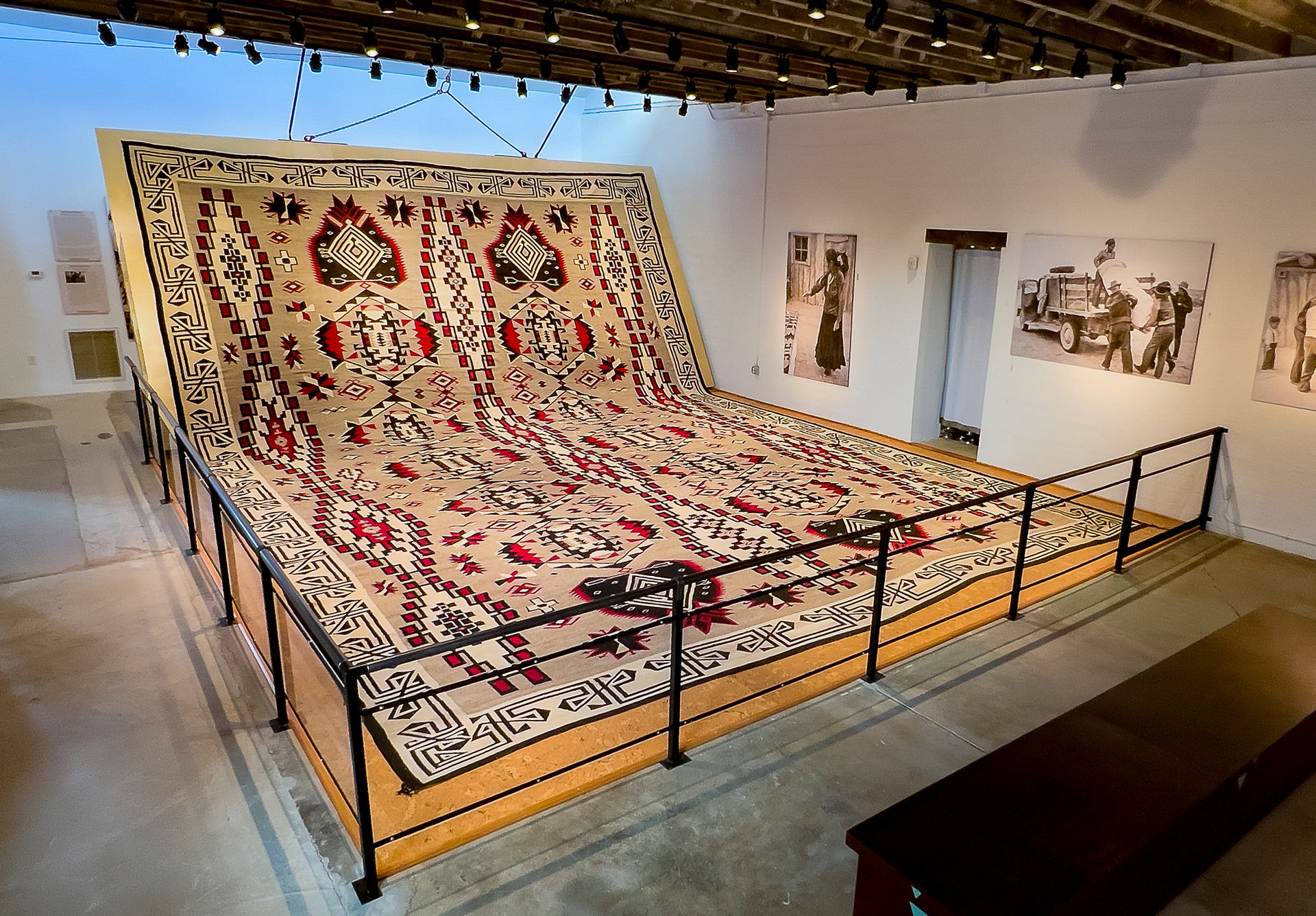 Large, intricately patterned rug on display in a gallery, raised on a wooden platform with a black railing.
