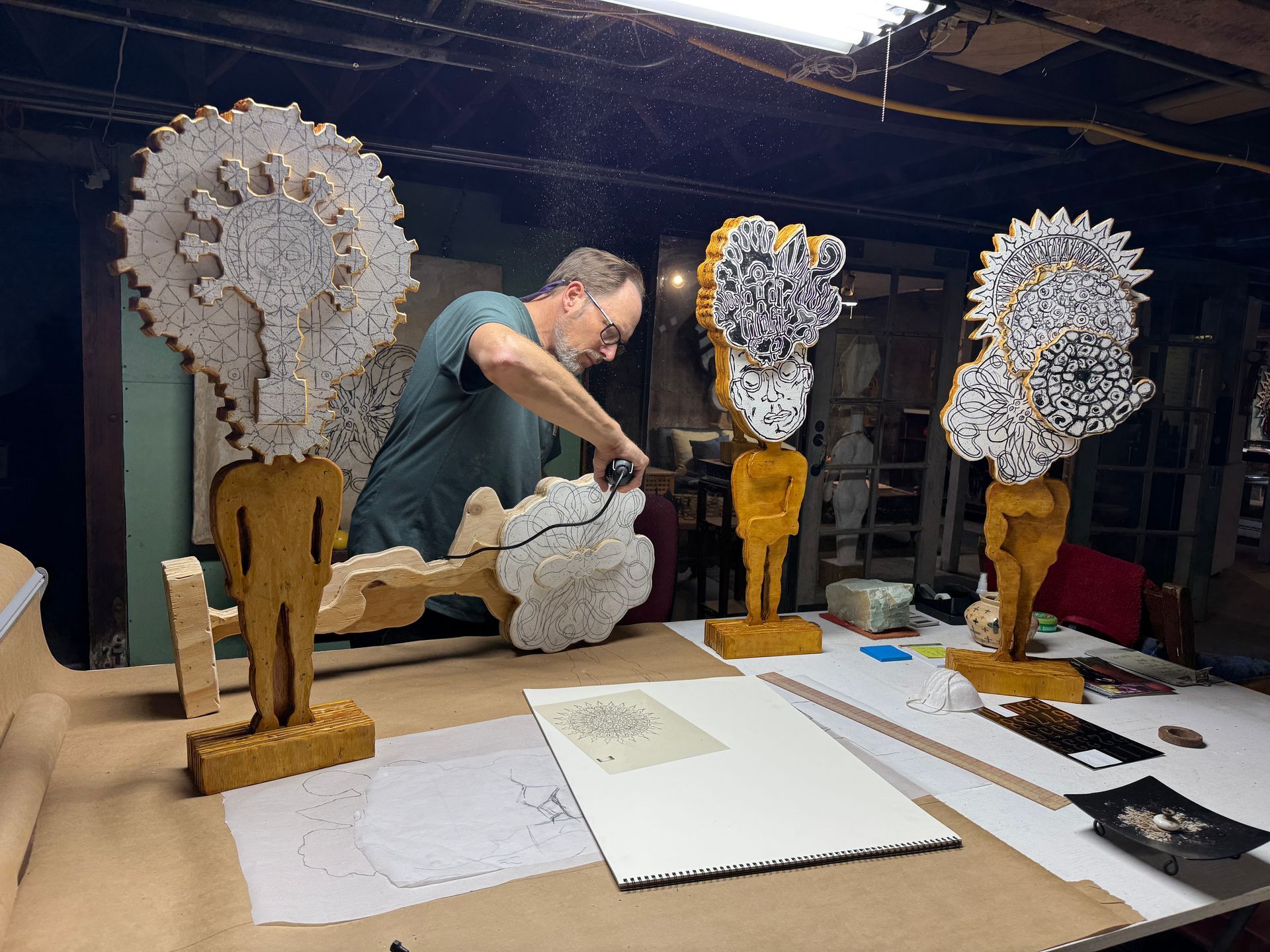 Man sitting at a table working on a wooden sculpture with several more sculptures around him.