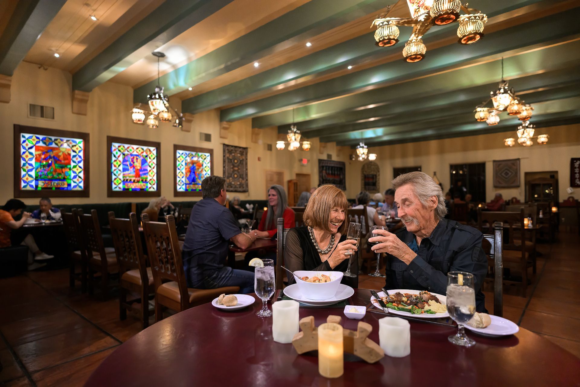 Couple toasting at a restaurant, red table with food and candles, stained glass, people dining in background.