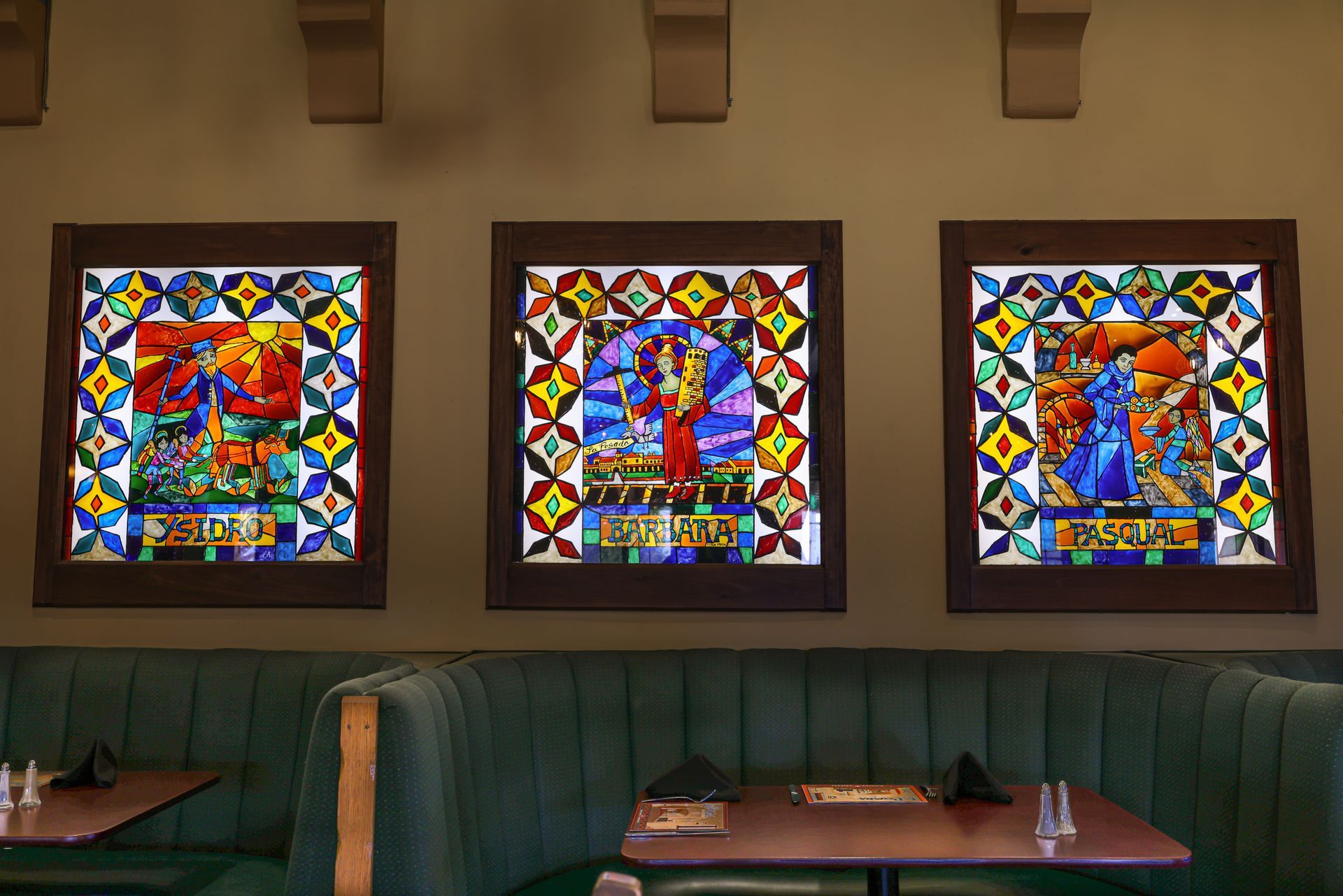 Three stained glass art panels on a wall above a green booth. Each panel depicts a figure with colorful designs.