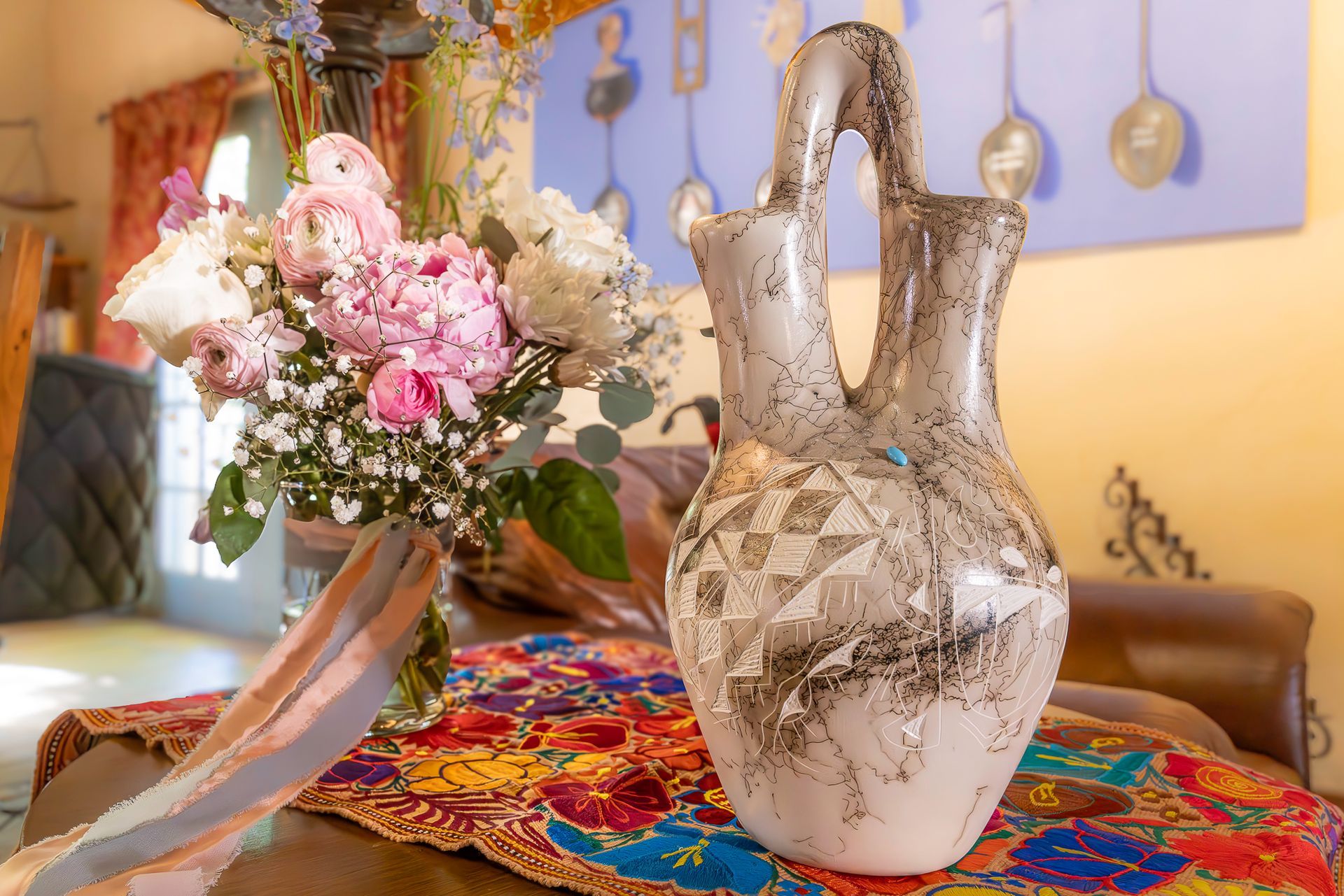 Vase with decorative floral design on a colorful embroidered table, flowers to the left, indoor setting.