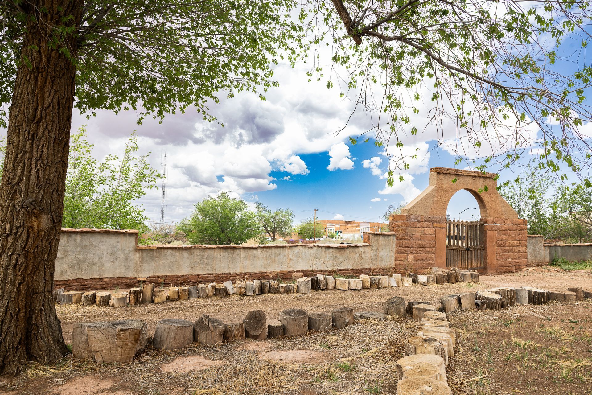 Old brick cemetery gate with stone fence, under a tree, against a cloudy blue sky.