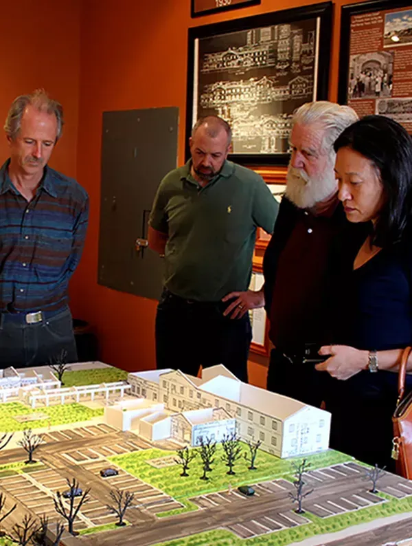 Four people viewing a building model; an orange-walled room with framed photos.