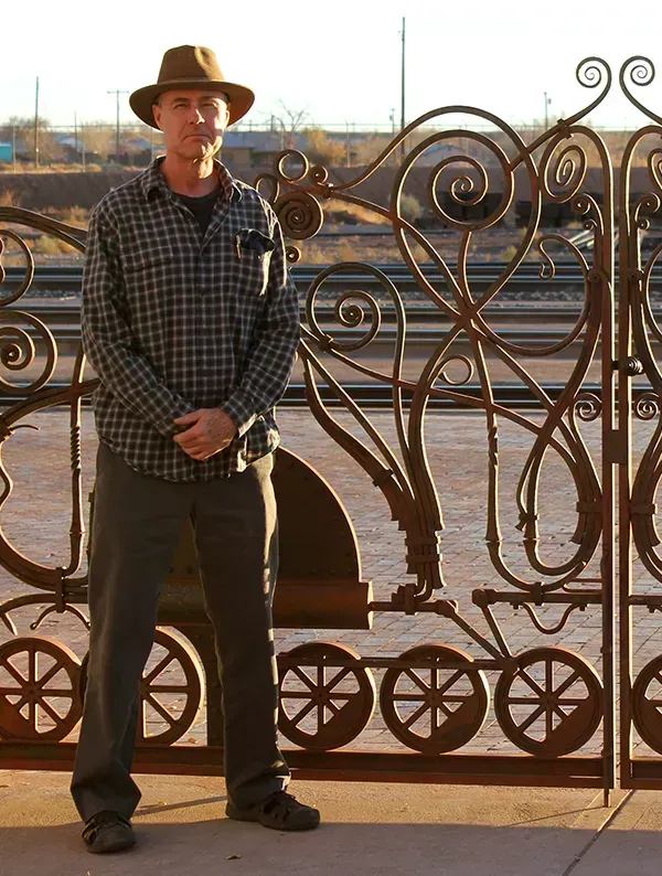 Man in hat stands beside a decorative metal gate in a railroad setting.