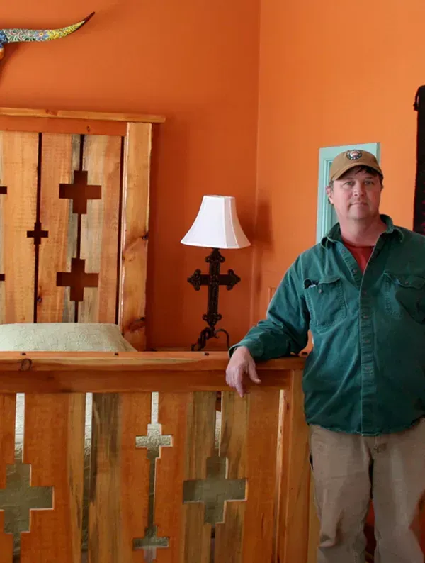 Man standing near a wooden bed with a cross design, against an orange wall.