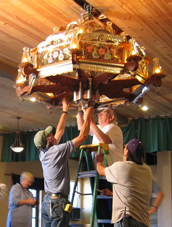 Four people installing a large, ornate chandelier indoors; some on a ladder, working at the fixture.