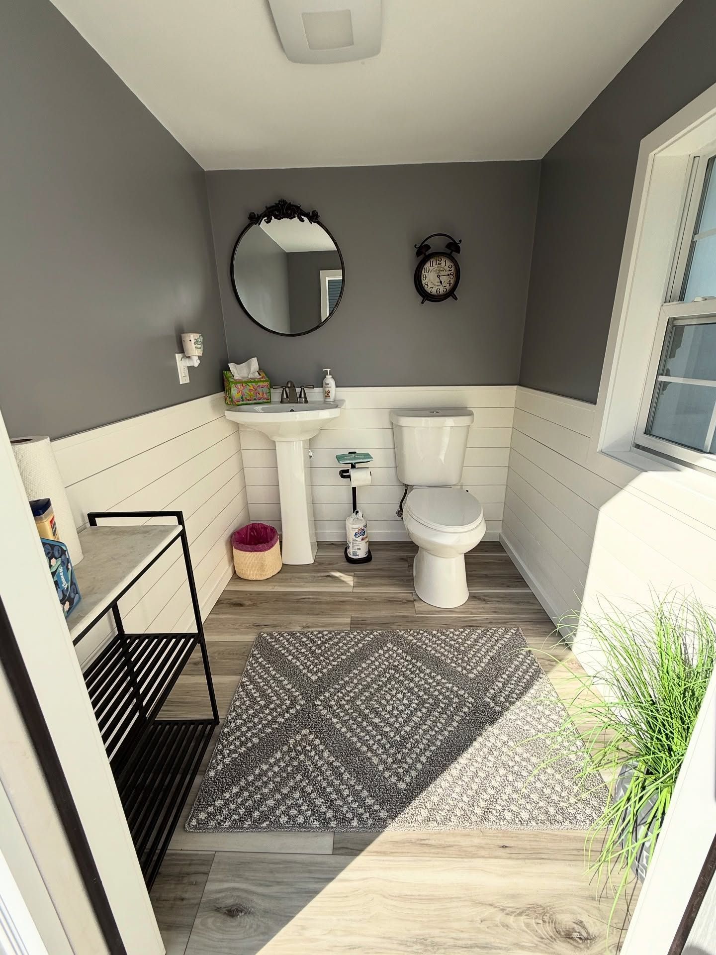 Small bathroom with gray walls, white wainscoting, pedestal sink, toilet, and a patterned rug.