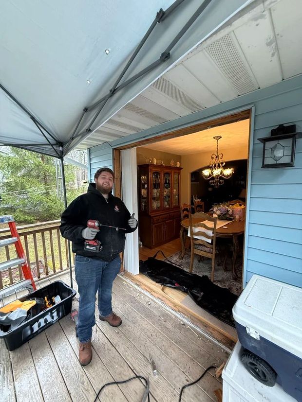 Man with a drill standing in a doorway, next to tools and a cooler, under a canopy on a porch.