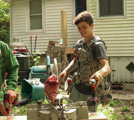 Young man smiling, using a drill outdoors. Wearing tool vest, near a saw and wood.