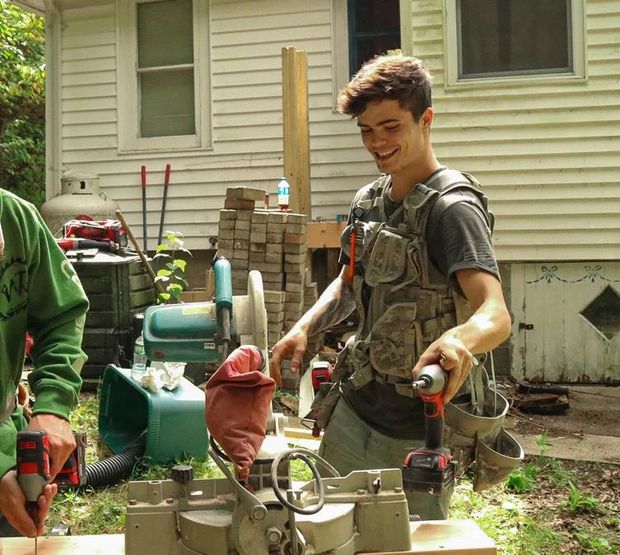 Young man smiling, using a drill outdoors. Wearing tool vest, near a saw and wood.