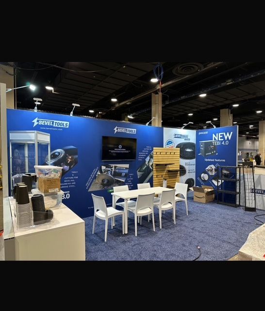Trade show booth with a blue backdrop displaying product images and a table with chairs.