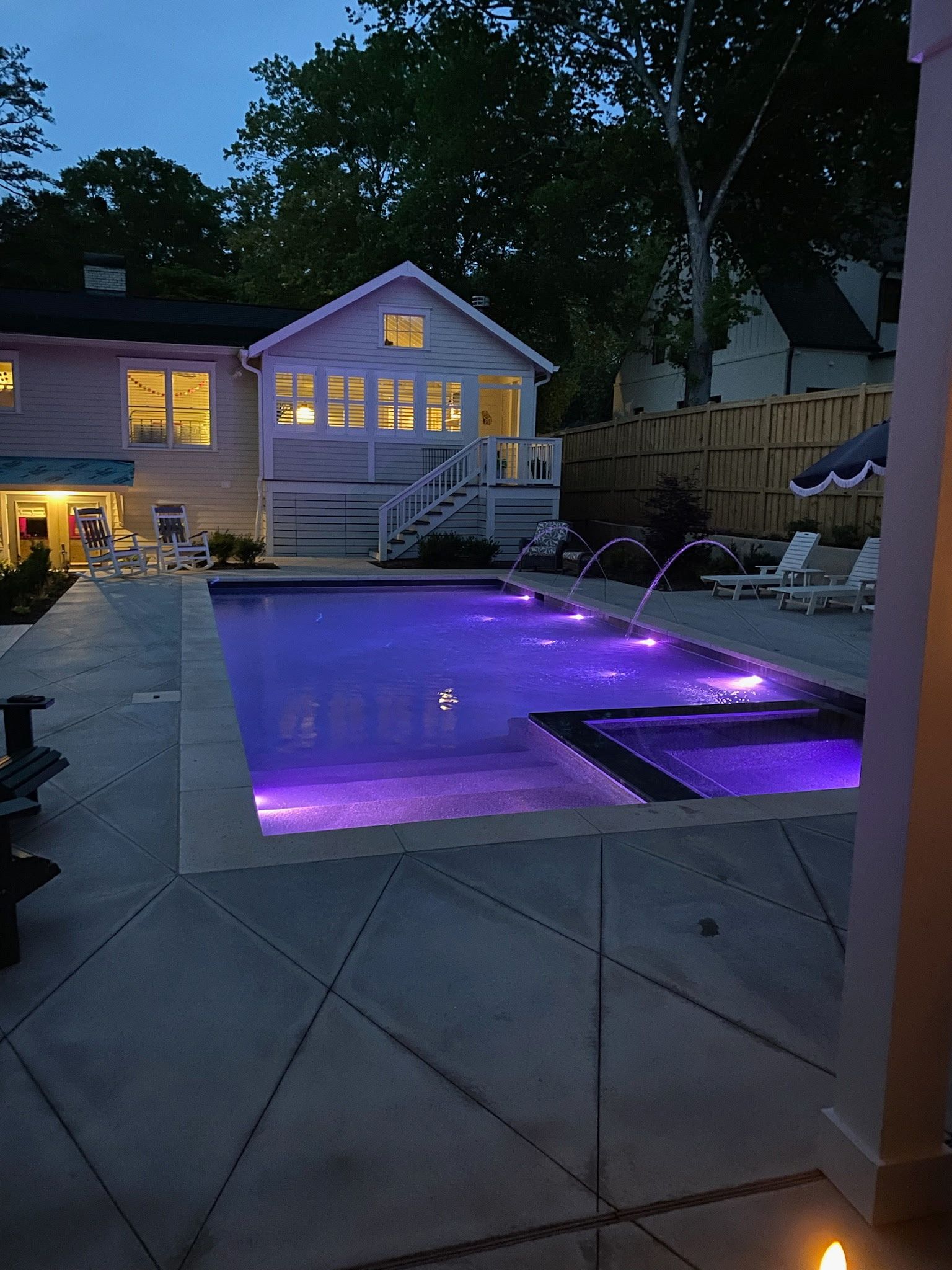 A backyard pool and attached spa illuminated in purple at dusk, with water jets, a patio, and a lit house in the background.