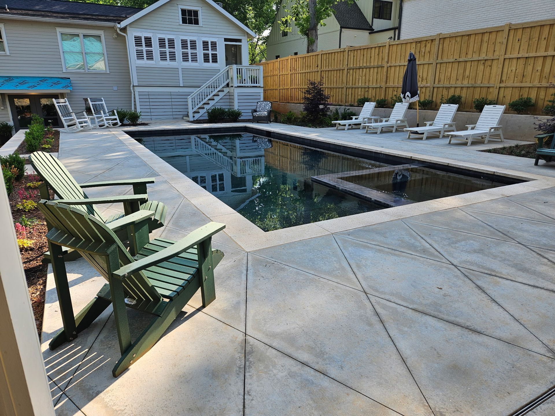A rectangular backyard pool with a built-in spa, surrounded by a concrete patio, lounge chairs, and two green chairs.