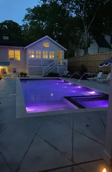A backyard pool and attached spa illuminated in purple at dusk, with water jets, a patio, and a lit house in the background.