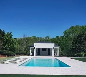 A rectangular swimming pool in front of a white pool house, surrounded by light stone decking and trees under a blue sky.