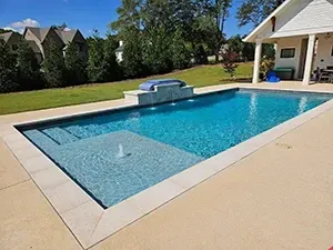 Rectangular swimming pool with blue water, a tanning ledge, fountain, and stone coping on a sunny day.