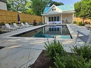 A backyard pool with lounge chairs on a patio, facing a small white house with a blue roof and a wooden fence.