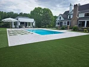 A backyard with a rectangular pool, a white covered patio, a striped umbrella, and a large house with a brick chimney.