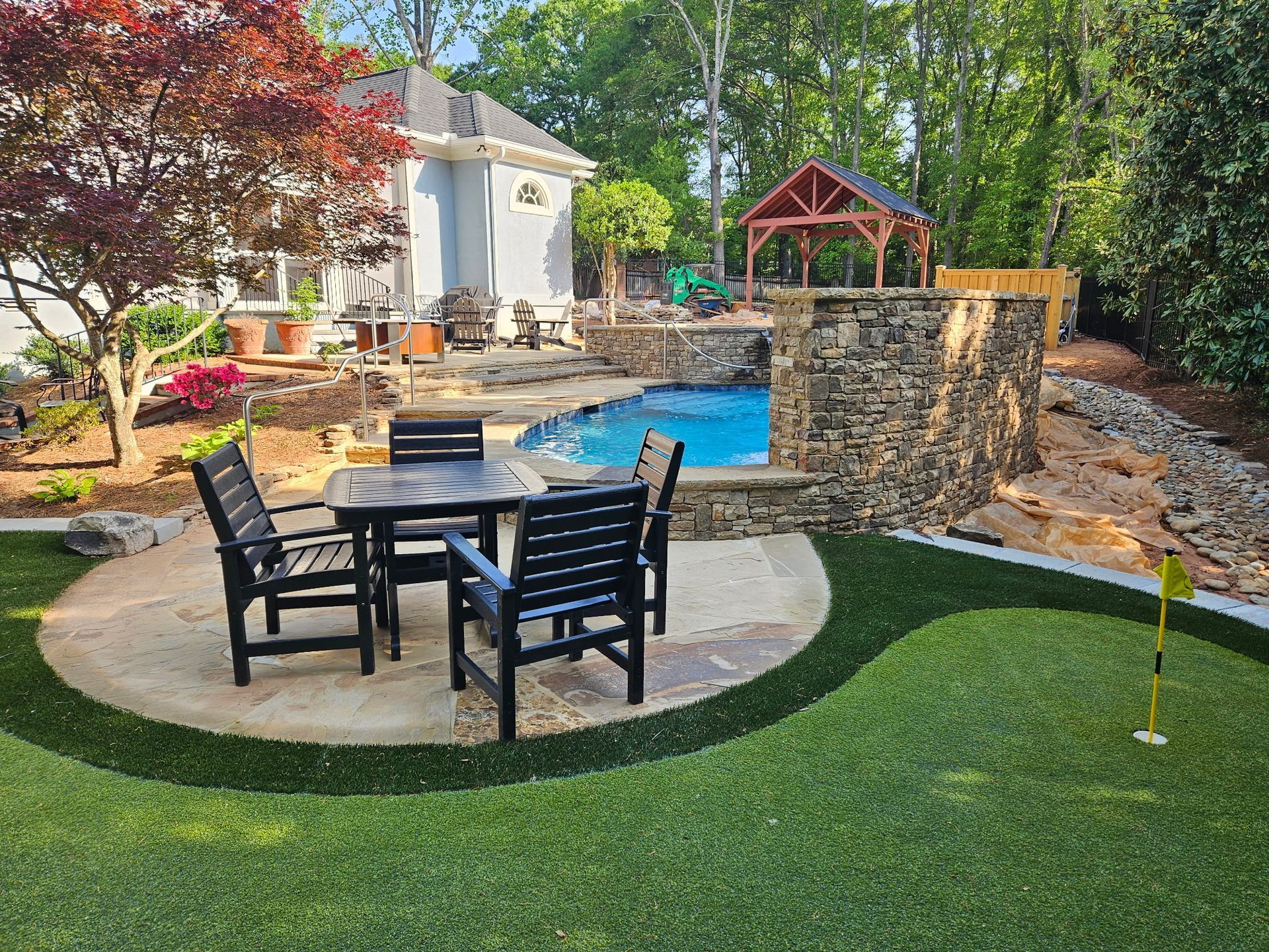 A backyard patio with a dining set on stone pavers, a small pool with a stone wall, and a putting green with a flag.