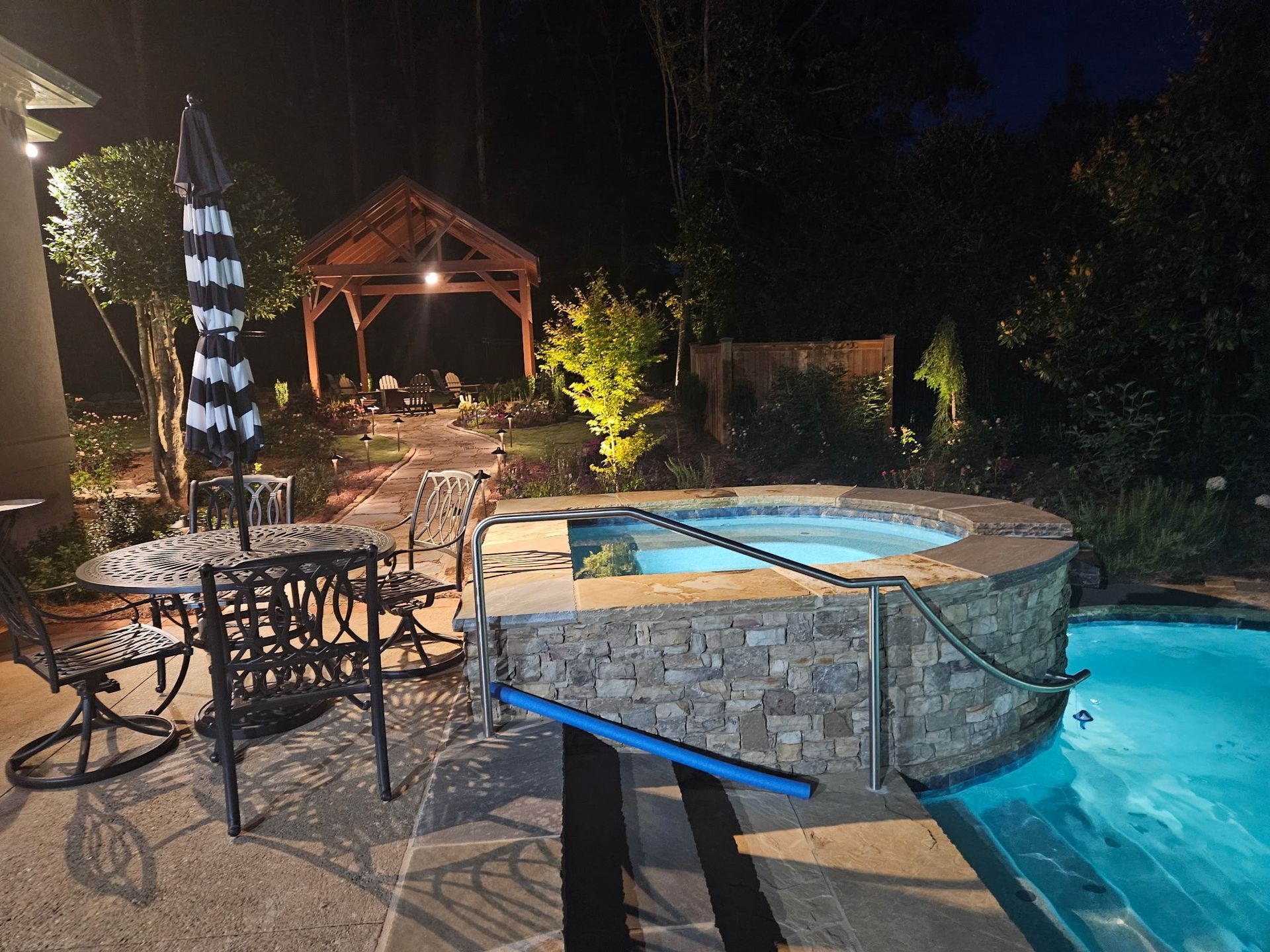 A backyard scene at night featuring an illuminated hot tub, an adjoining swimming pool, patio furniture, and a gazebo.