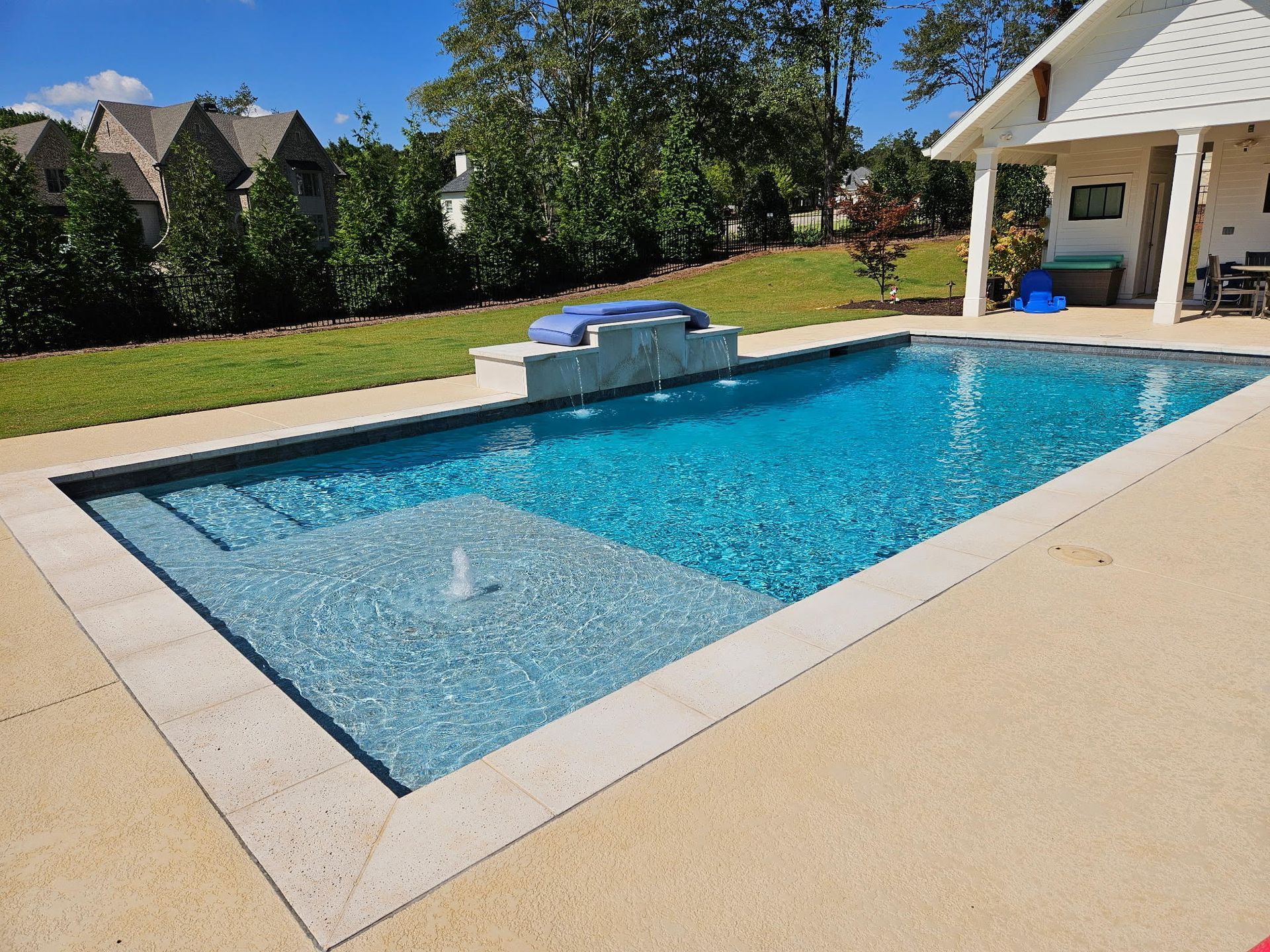 A rectangular backyard pool with a sun ledge, fountain, and stone coping, surrounded by a light-colored concrete deck.