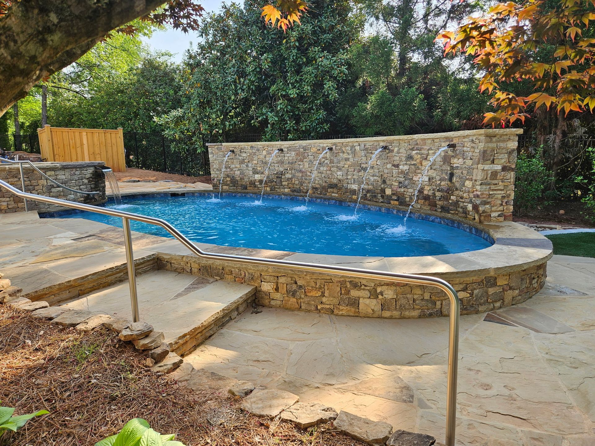 A stone-walled, oval backyard pool with a built-in water feature, surrounded by paved decking and lush trees.
