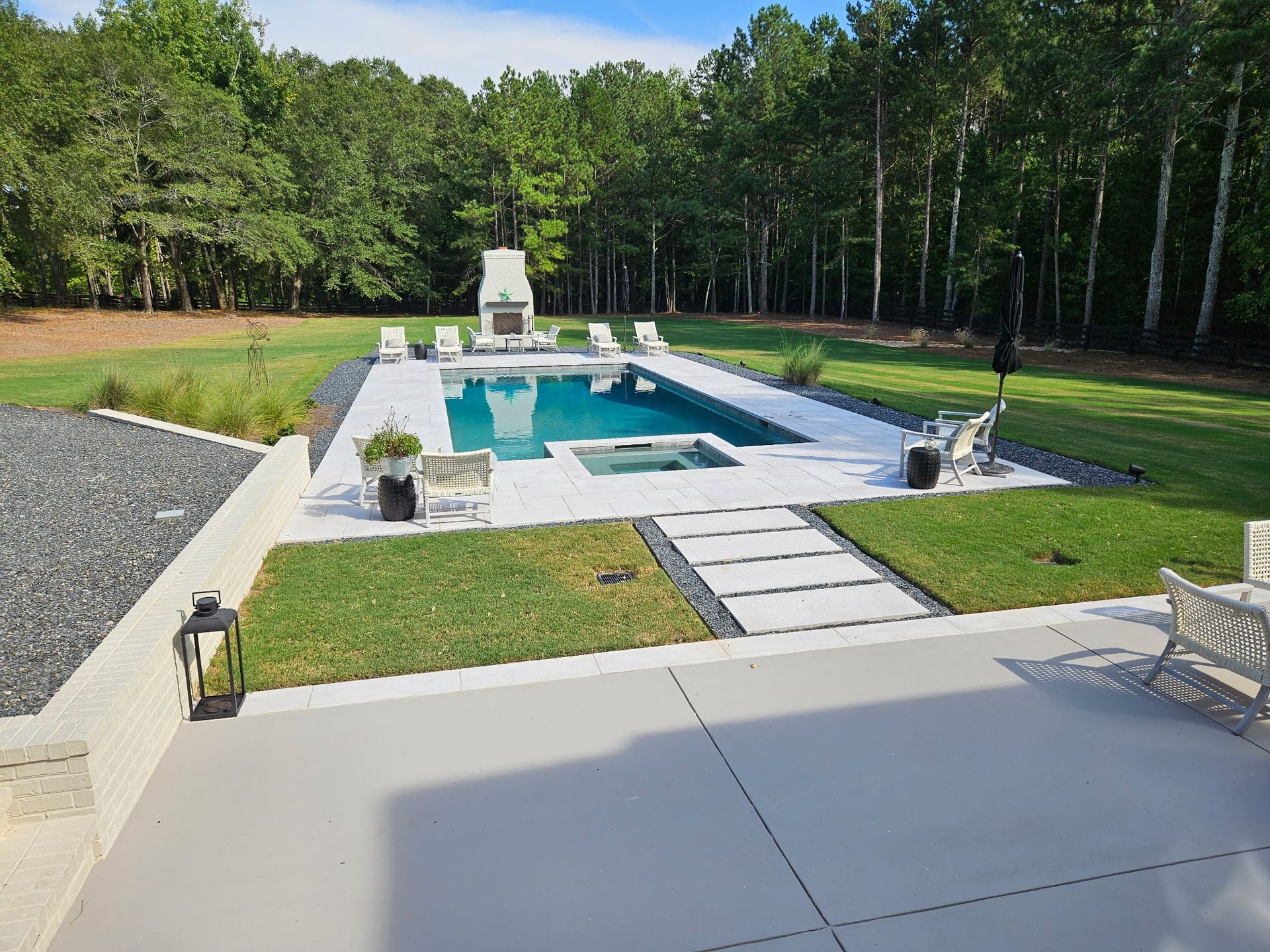 A rectangular swimming pool with a hot tub and stone fireplace set in a spacious backyard bordered by trees.