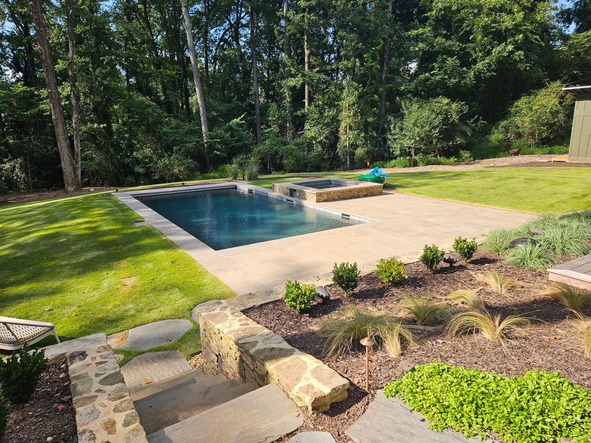 A backyard with a rectangular swimming pool, stone patio, stone steps, and landscaping next to a dense forest.